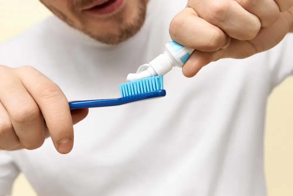 A person in a white shirt squeezing toothpaste from a tube onto a blue toothbrush.
