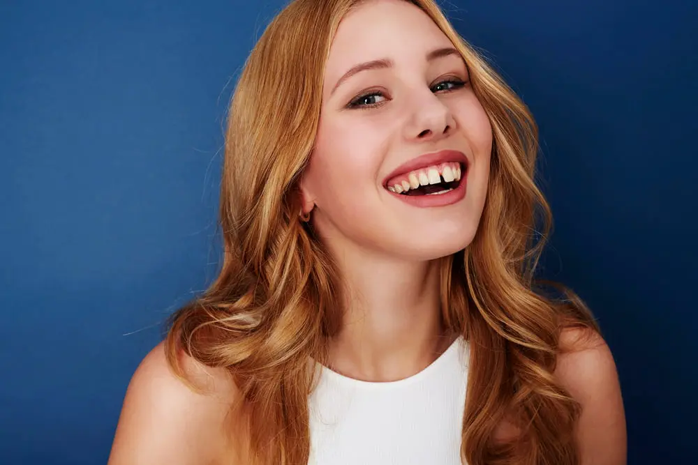 A young woman with long, wavy blonde hair smiles broadly, wearing a white sleeveless top against a blue background.