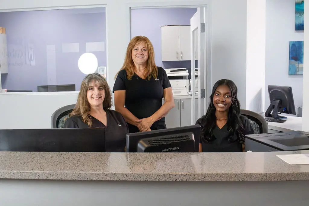 Three women in black tops are behind a reception desk in an office setting. Two are seated at computer monitors, and one is standing behind them.