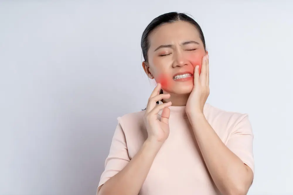 A person winces in pain, holding their hand to the side of their face with a visible red area on their cheek, indicating a toothache or jaw pain.