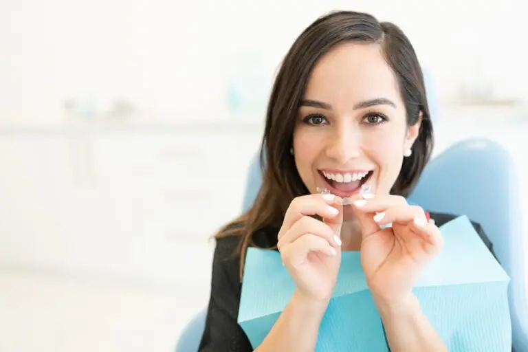 A woman sits in a dental chair holding two clear aligners, smiling, with a dental bib around her neck.
