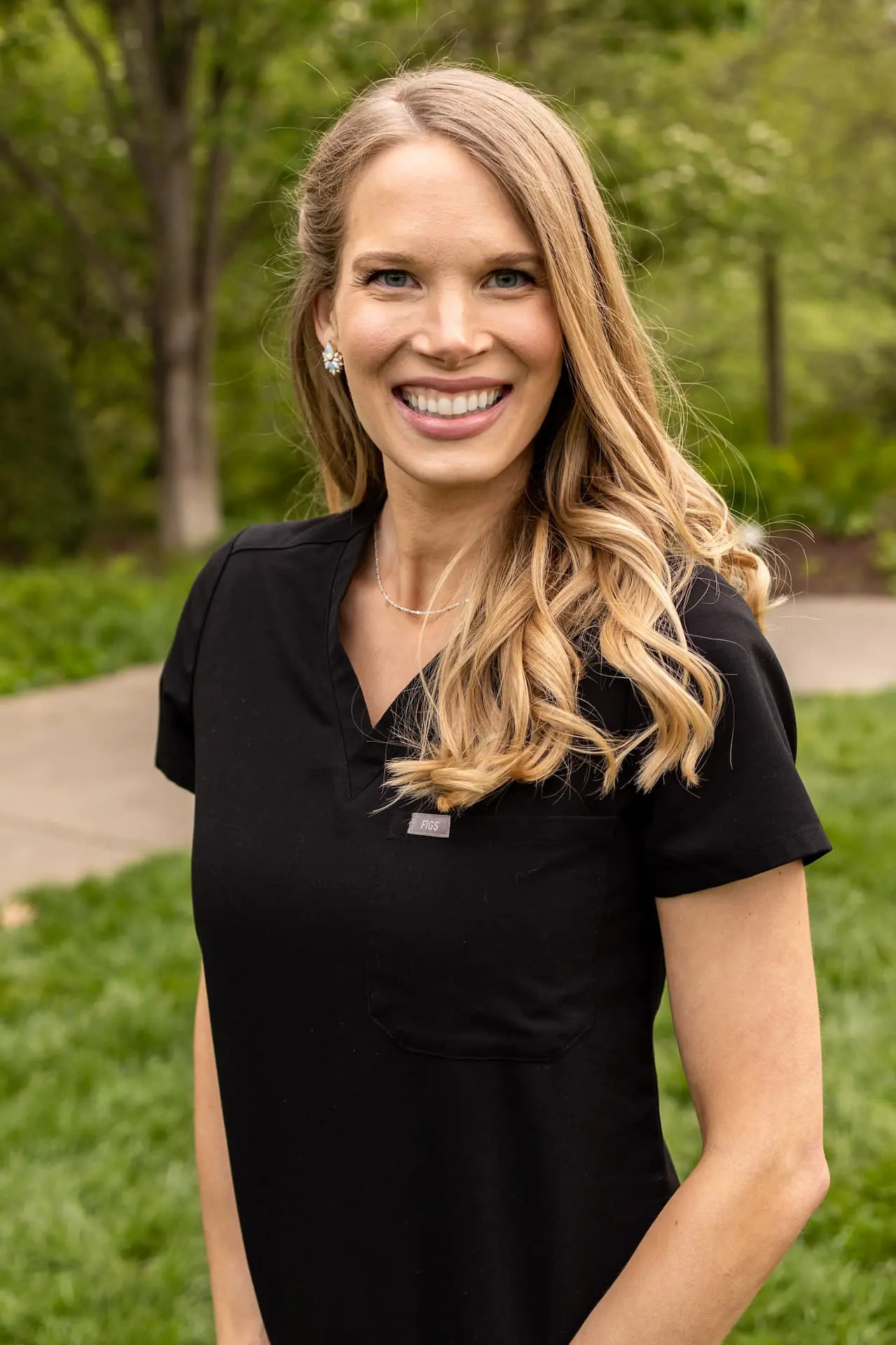 Dr. Nicole Crasto smiles warmly outdoors, wearing a black shirt with a name tag. The serene backdrop of trees and grass adds a refreshing touch to the scene in Liberty Township or Hamilton, OH