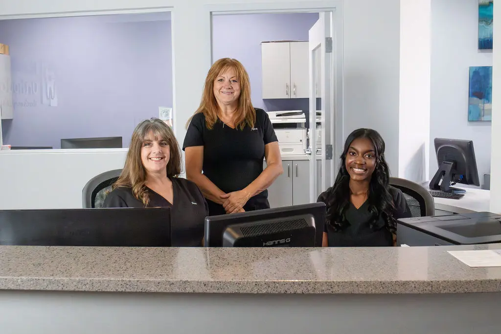 Three smiling women, possibly part of the team at Queen City Orthodontics in Liberty Township and Hamilton, OH, pose behind a reception desk with computer monitors, suggesting career opportunities at the clinic.