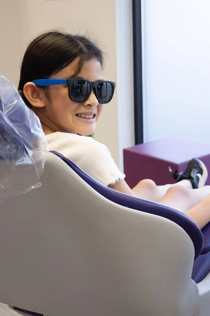 A child wearing sunglasses and braces smiles while sitting in a dental chair.