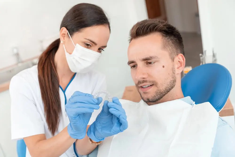 A dentist shows a male patient an orthodontic aligner during a dental consultation. The patient is seated in a dental chair wearing a bib, while the dentist wears gloves and a mask at Queen City Orthodontics in Liberty Township and Hamilton OH.