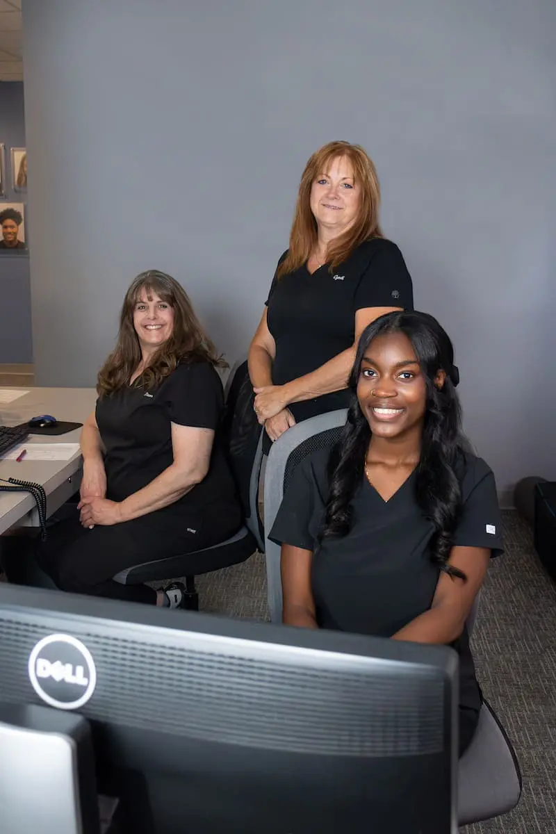Three women wearing black uniforms are in an office setting. One woman is seated at a desk with a computer, another is seated beside her, and the third is standing behind them at Queen City Orthodontics in Liberty Township or Hamilton, OH.
