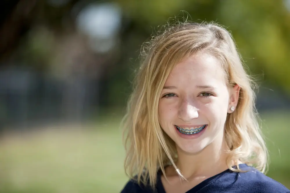 A young person with shoulder-length blonde hair and braces is smiling outdoors against a blurred green background.