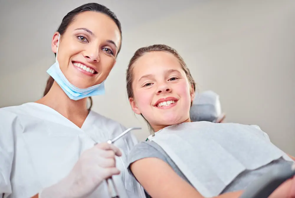 A dental professional in white attire and face mask lowers is standing next to a smiling young girl in a dentist's chair, holding a dental instrument.