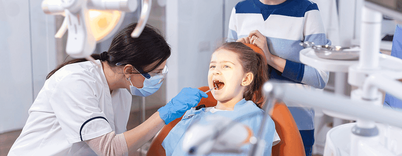 A dentist at Queen City Orthodontics examines a young girl’s teeth in a dental office. The girl is seated comfortably in an orange chair, with an adult standing nearby. Various dental equipment surrounds them, ensuring the best care for her vibrant smile in Liberty Township or Hamilton, OH
