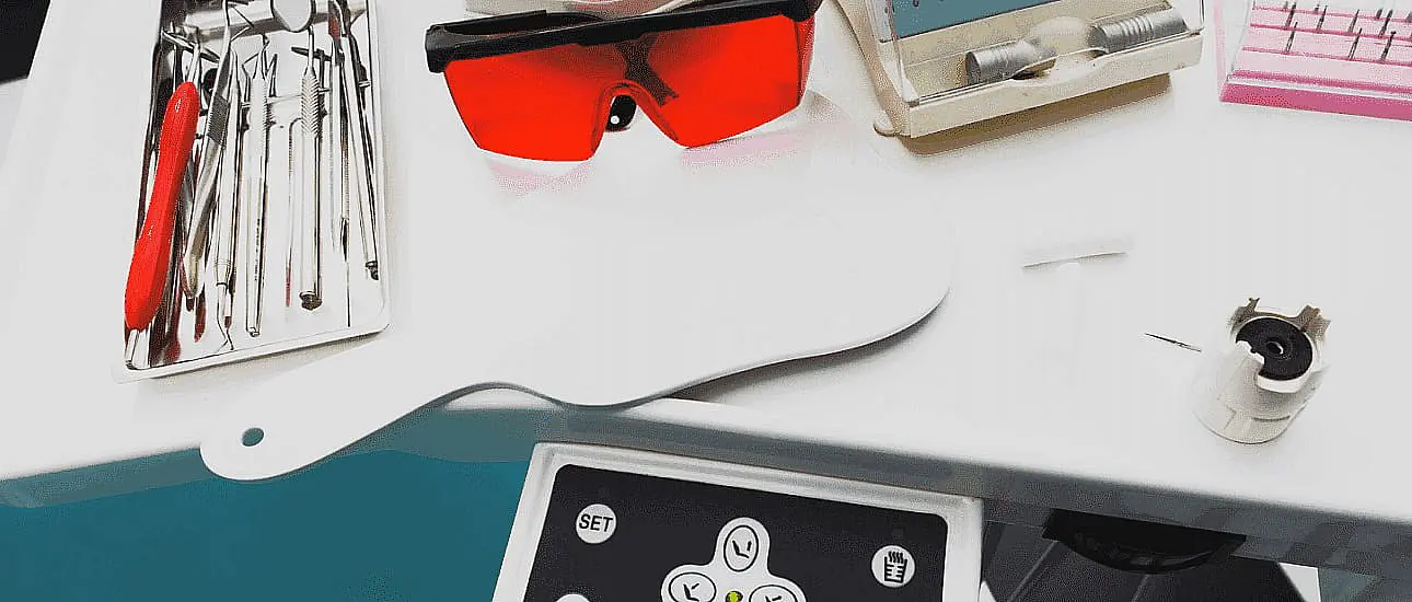 Dental tools, metal braces, and protective red glasses rest on a dental tray, alongside a control panel and equipment on a teal surface in Liberty Township or Hamilton, OH