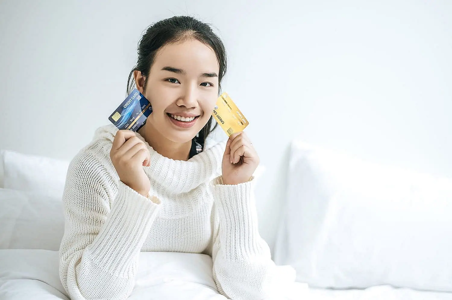 Smiling person in a white sweater, possibly considering a braces payment plan, holds a blue credit card in one hand and a gold credit card in the other while sitting on a white bed in Liberty Township or Hamilton, OH