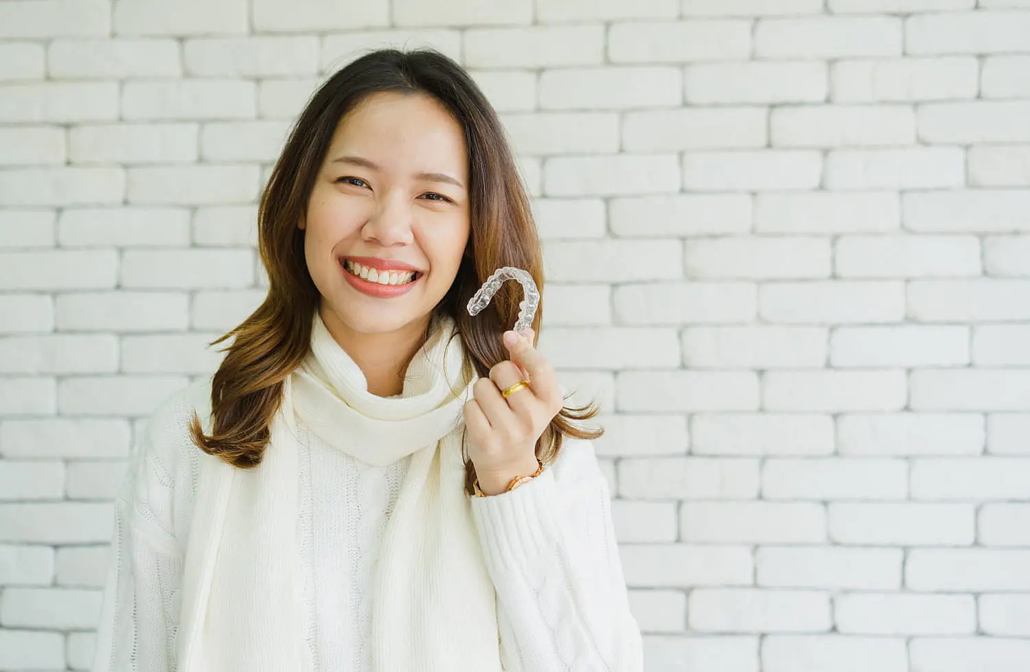 A woman in a white sweater and scarf smiles while proudly displaying her Invisalign® Clear Aligners against a white brick wall in Liberty Township, OH
