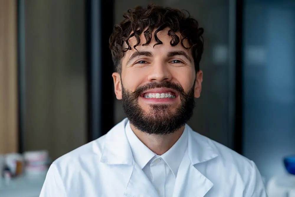 A smiling man with curly hair and a beard, sporting clear ceramic braces, stands indoors wearing a white lab coat in Liberty Township, OH