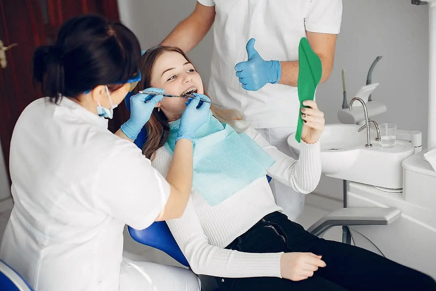 A dental patient in a chair holds a mirror while a female dentist adjusts orthodontic appliances. An assistant stands nearby holding tools in Liberty Township, OH