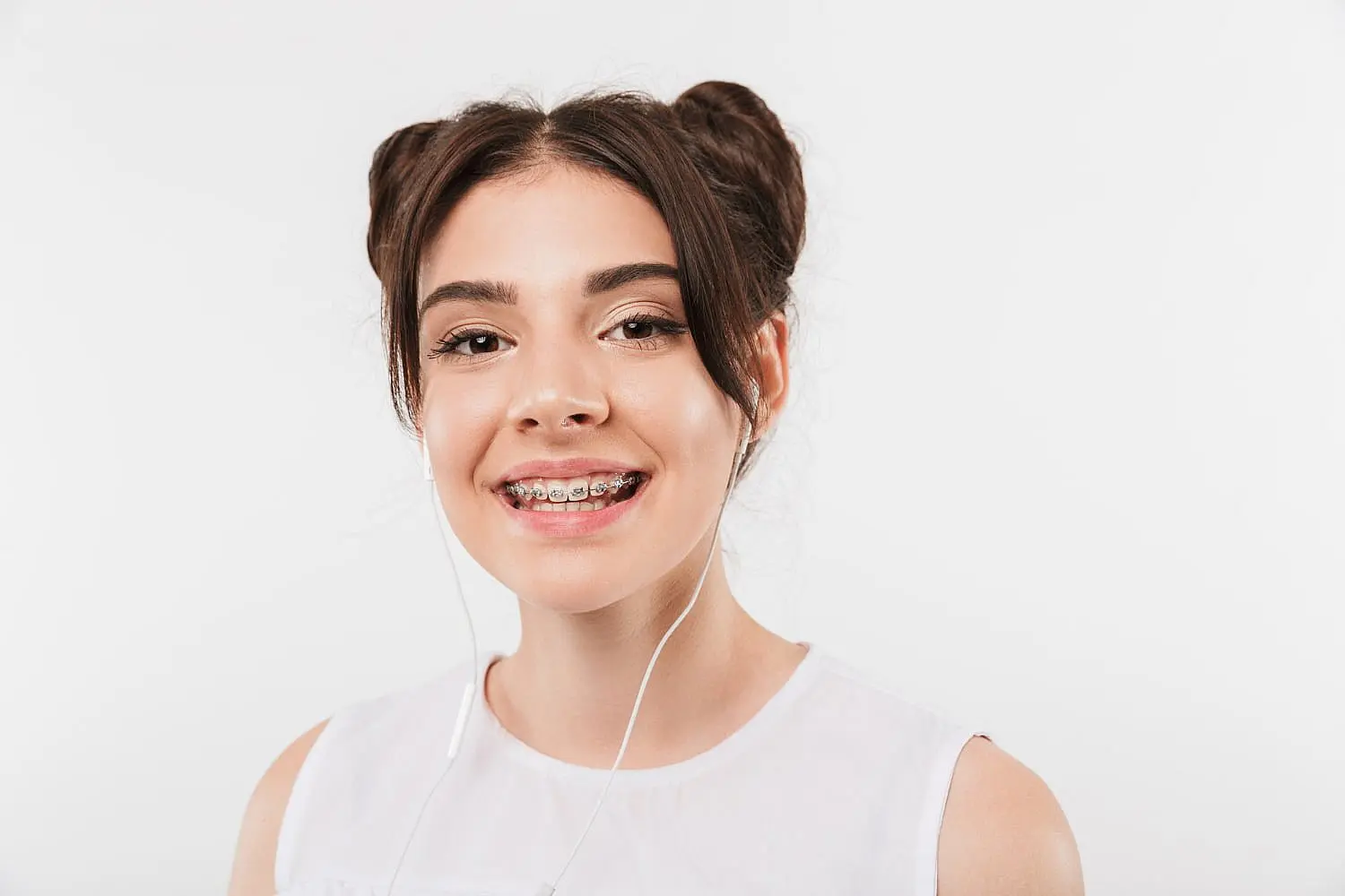 A young woman with braided hair and metal braces smiles while wearing earbuds. She is dressed in a white top against a plain background in Liberty Township or Hamilton, OH