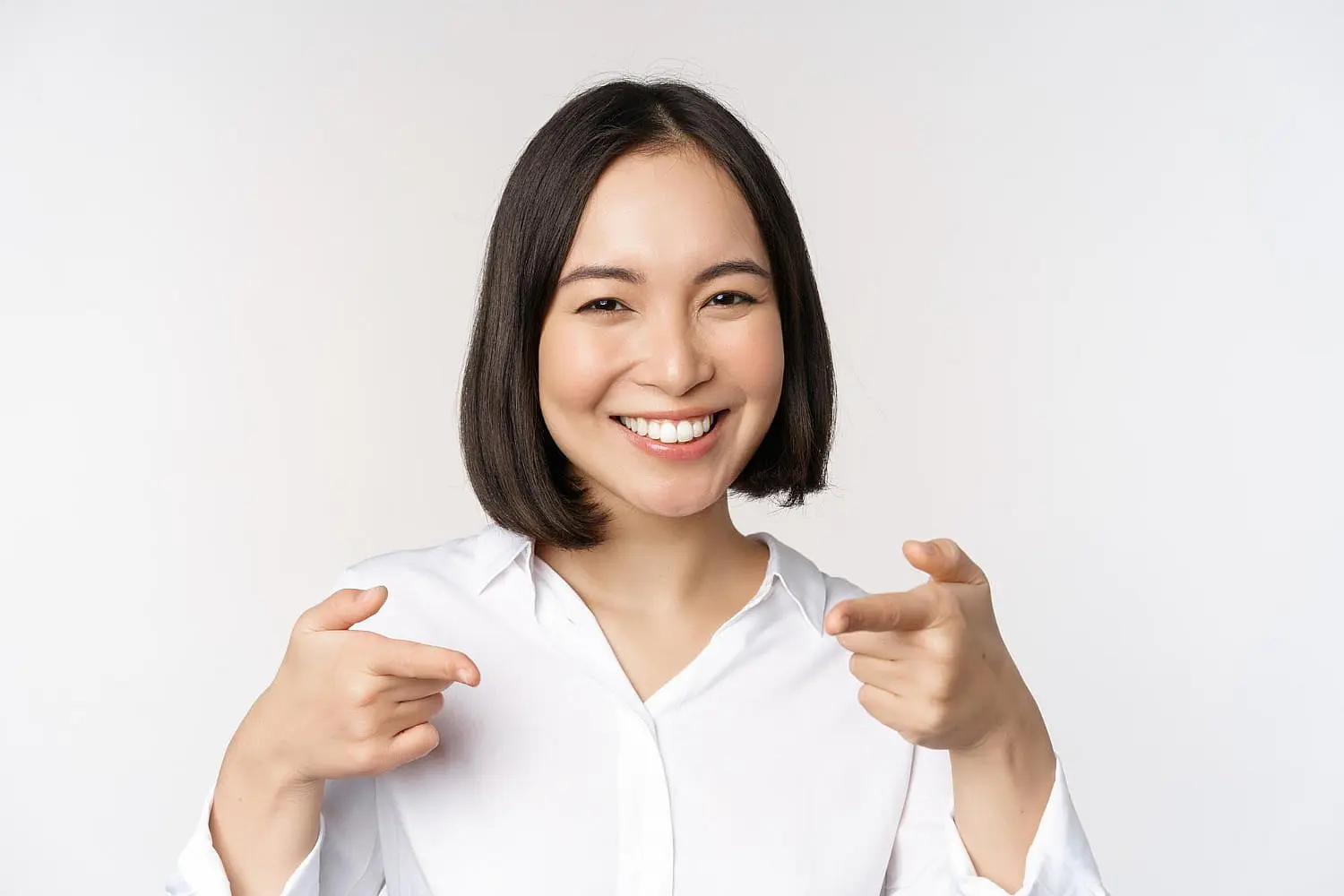 Smiling person with short dark hair in a white shirt, pointing fingers towards the camera against a plain white background at Queen City Orthodontics in Liberty Township and Hamilton OH.
