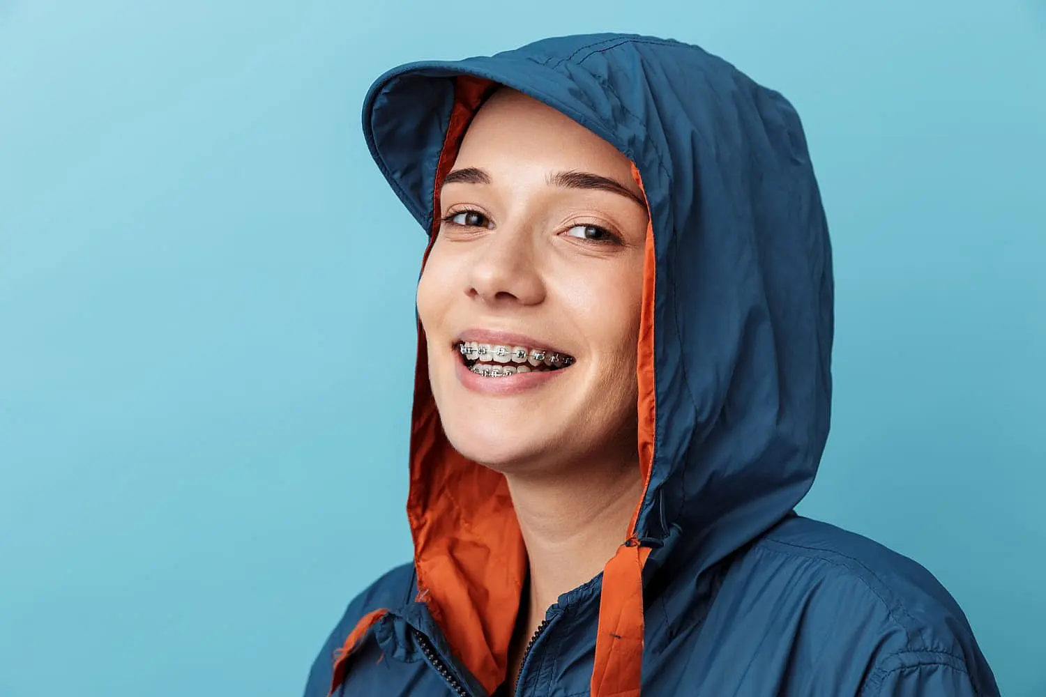 A person in a blue jacket with an orange hood flashes a bright smile, proudly displaying their braces against the blue background in Liberty Township, OH