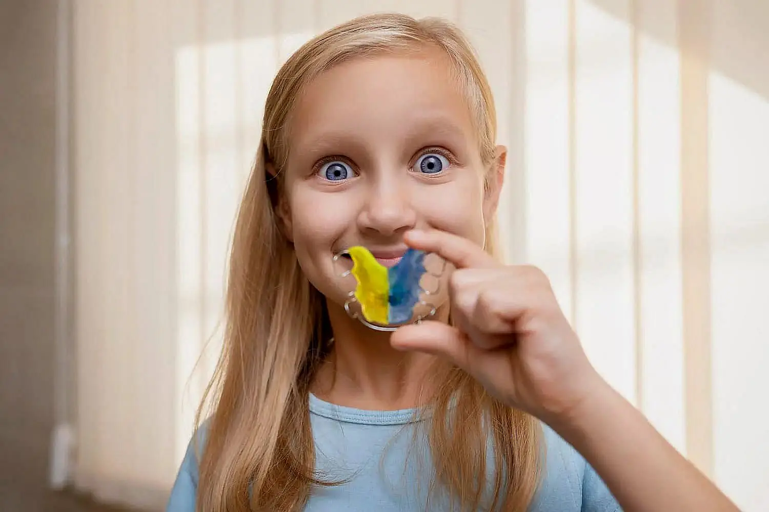 A girl with long blonde hair holds a colorful orthodontic appliance, smiling and showing it towards the camera in a well-lit room in Liberty Township, OH