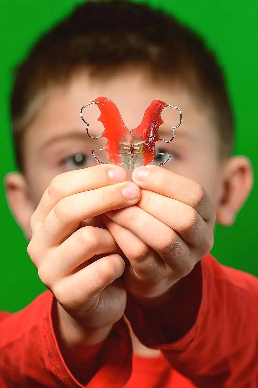 A child carefully holds a red dental retainer, an essential orthodontic appliance, with both hands against a vibrant green background in Hamilton, OH