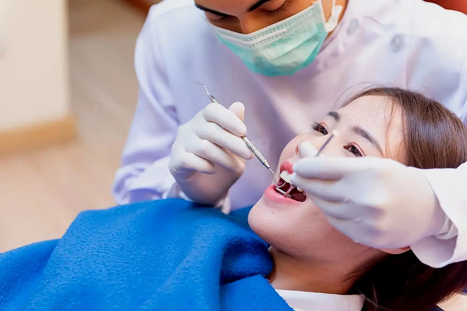 The dentist, masked and focused, examines the patient's teeth using dental tools. An array of orthodontic appliances glints in the background as the patient reclines under a blue blanket in Hamilton, OH
