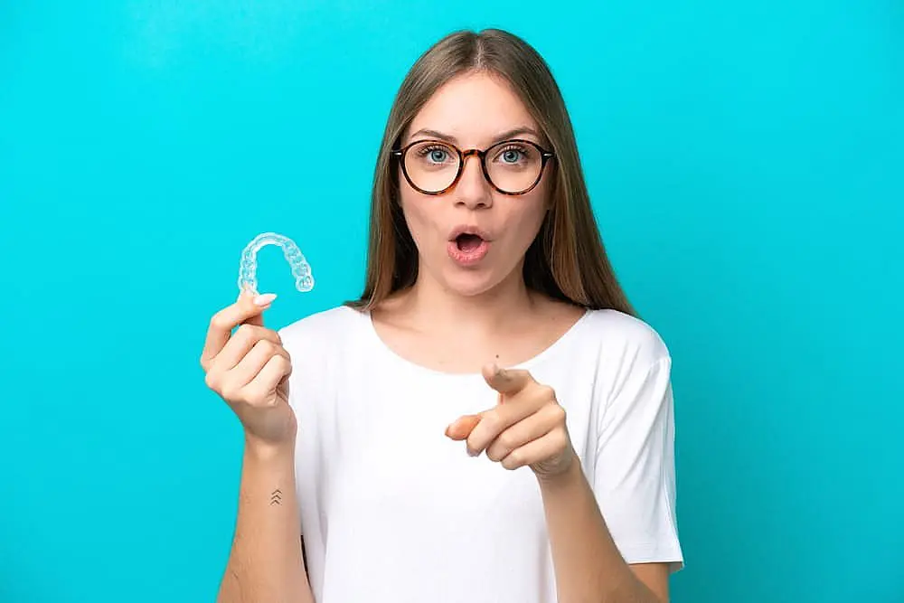 A woman with glasses holds Invisalign® Clear Aligners in one hand and points with the other against a blue background. She appears to be speaking or expressing surprise in Hamilton, OH