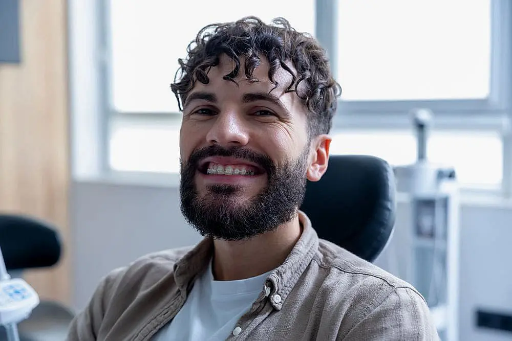 A person with a beard and curly hair is sitting in a chair, smiling and showing clear braces. There is a window in the background in Hamilton OH