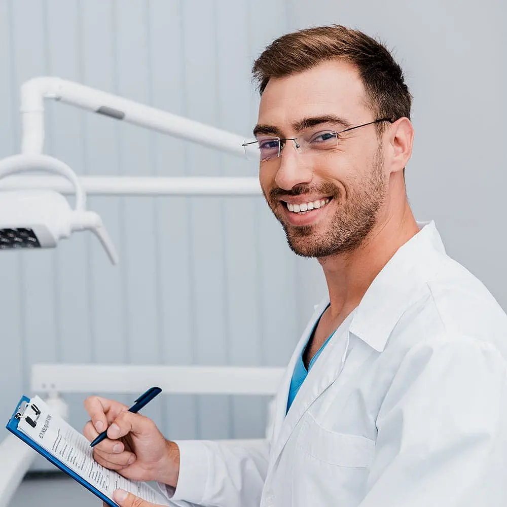 A man in a white coat holds a clipboard and pen, smiling while seated in a dental office in Liberty Township and Hamilton, OH.