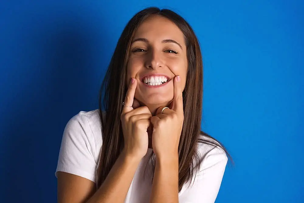 Person in a white shirt against a blue background, smiling and pointing at their cheeks with both index fingers in Liberty Township, OH