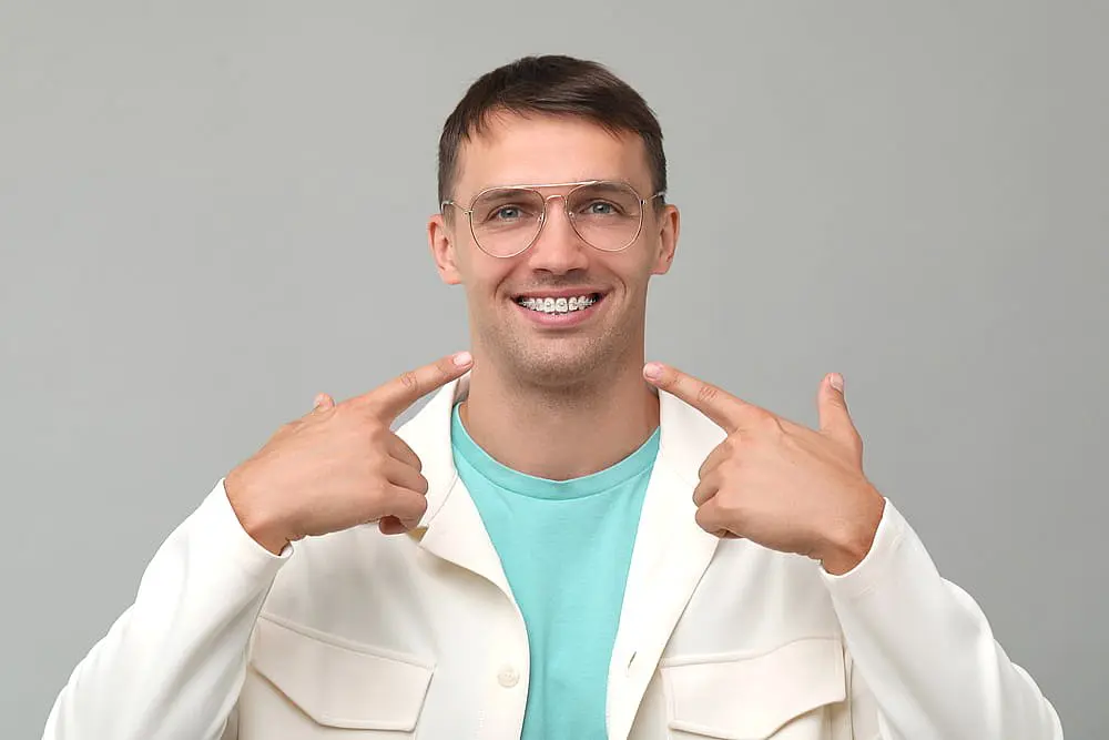 A man with braces and glasses smiles, proudly showcasing his teeth with both index fingers. Dressed in a white jacket over a turquoise shirt, he stands against a simple gray backdrop in Hamilton, OH