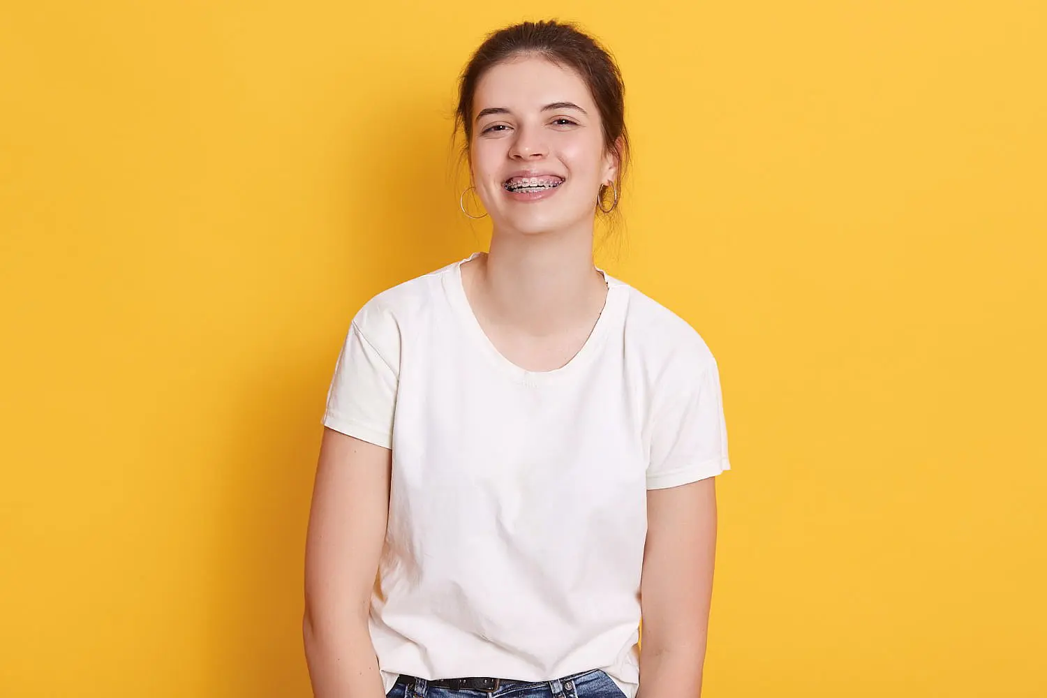 A smiling person with braces beams against a vibrant yellow background, showcasing their hoop earrings and crisp white t-shirt in Hamilton, OH