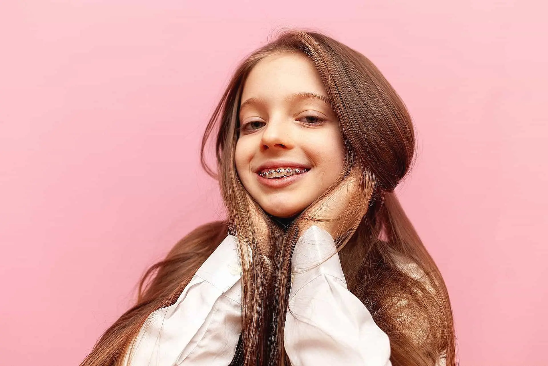 A young girl beams confidently, her braces glinting as she stands against a vivid pink background in a crisp white shirt in Liberty Township or Hamilton, OH