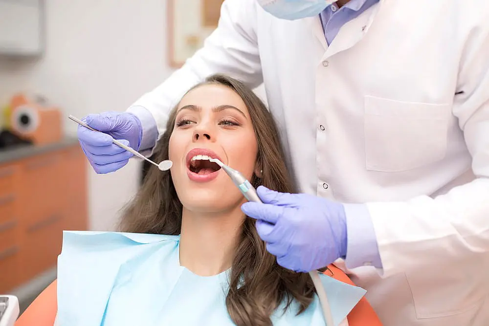 A dentist examines a woman's teeth, checking her braces with precision. She is seated in the clinic wearing a dental bib, while the dentist, clad in a white coat and protective gloves, skillfully uses dental tools for the assessment in Hamilton, OH