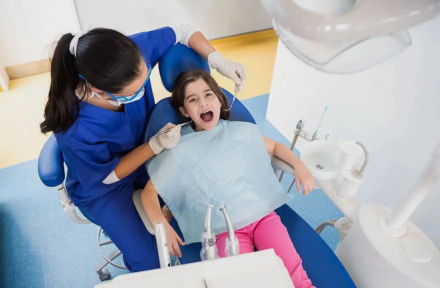 A dentist in blue scrubs carefully examines a young girl in a dental chair, surrounded by dental tools and orthodontic appliances. The girl, wearing a bib, has her mouth open wide, showcasing the attentive care she receives in Liberty Township, OH
