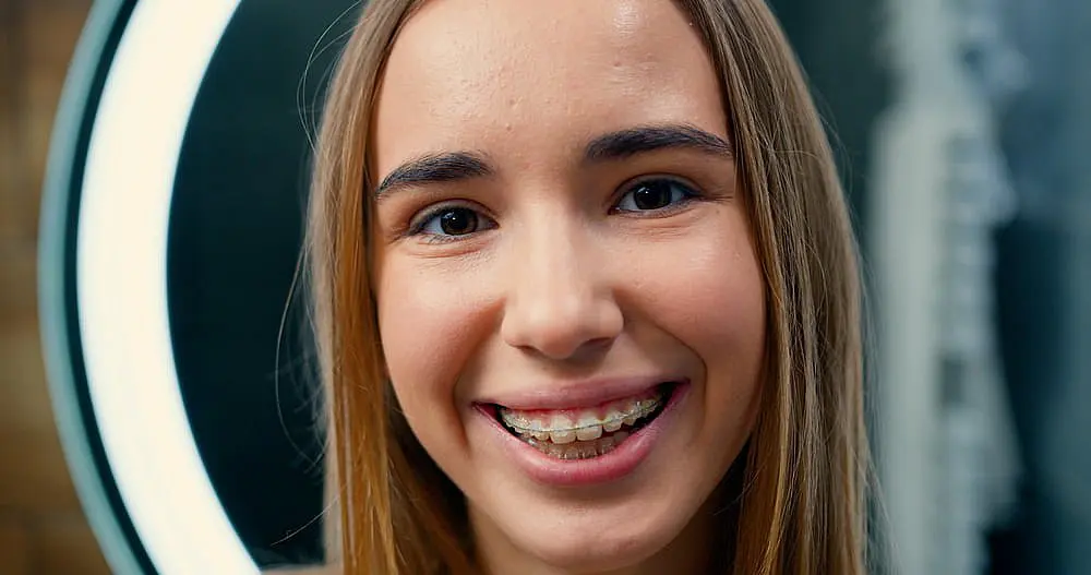 A smiling person with long hair and clear braces is illuminated by a ring light in the background, lending a soft glow to their joyful expression in Hamilton, OH