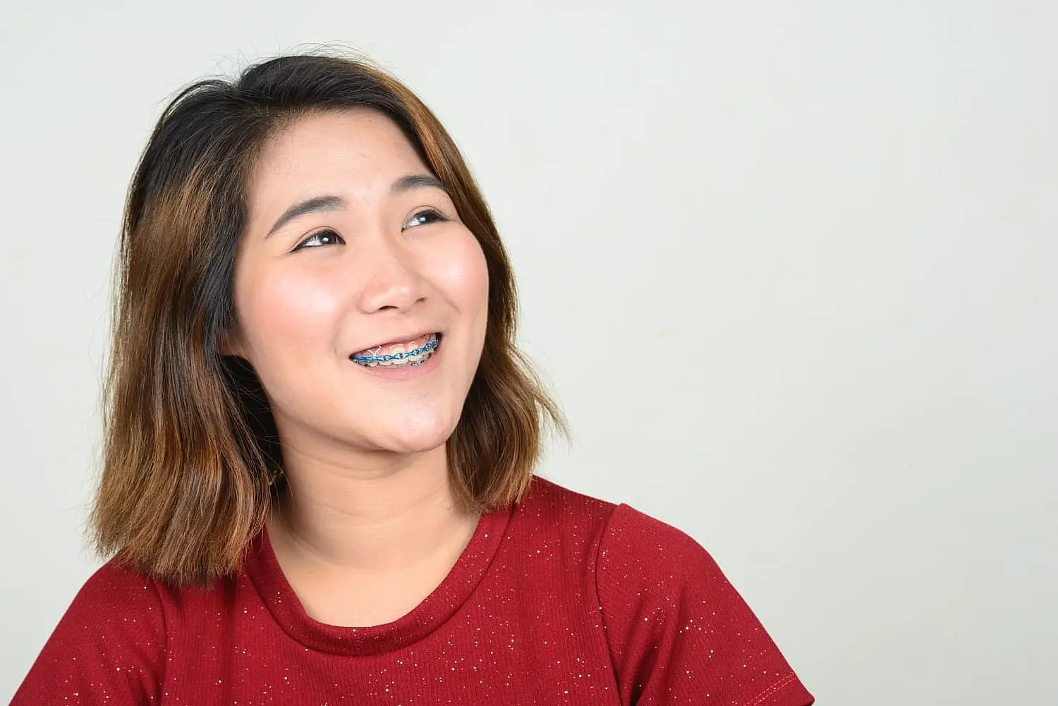 A person with shoulder-length hair beams, showcasing metal braces as they gaze upward, adorned in a red shirt against a plain background in Liberty Township or Hamilton, OH