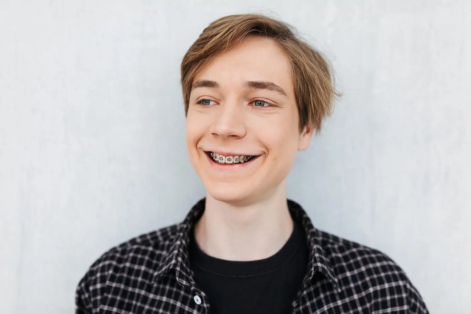 A smiling young person wearing a plaid shirt proudly showcases their dazzling smile, featuring stylish types of braces, as they stand against a light background in Liberty Township or Hamilton, OH