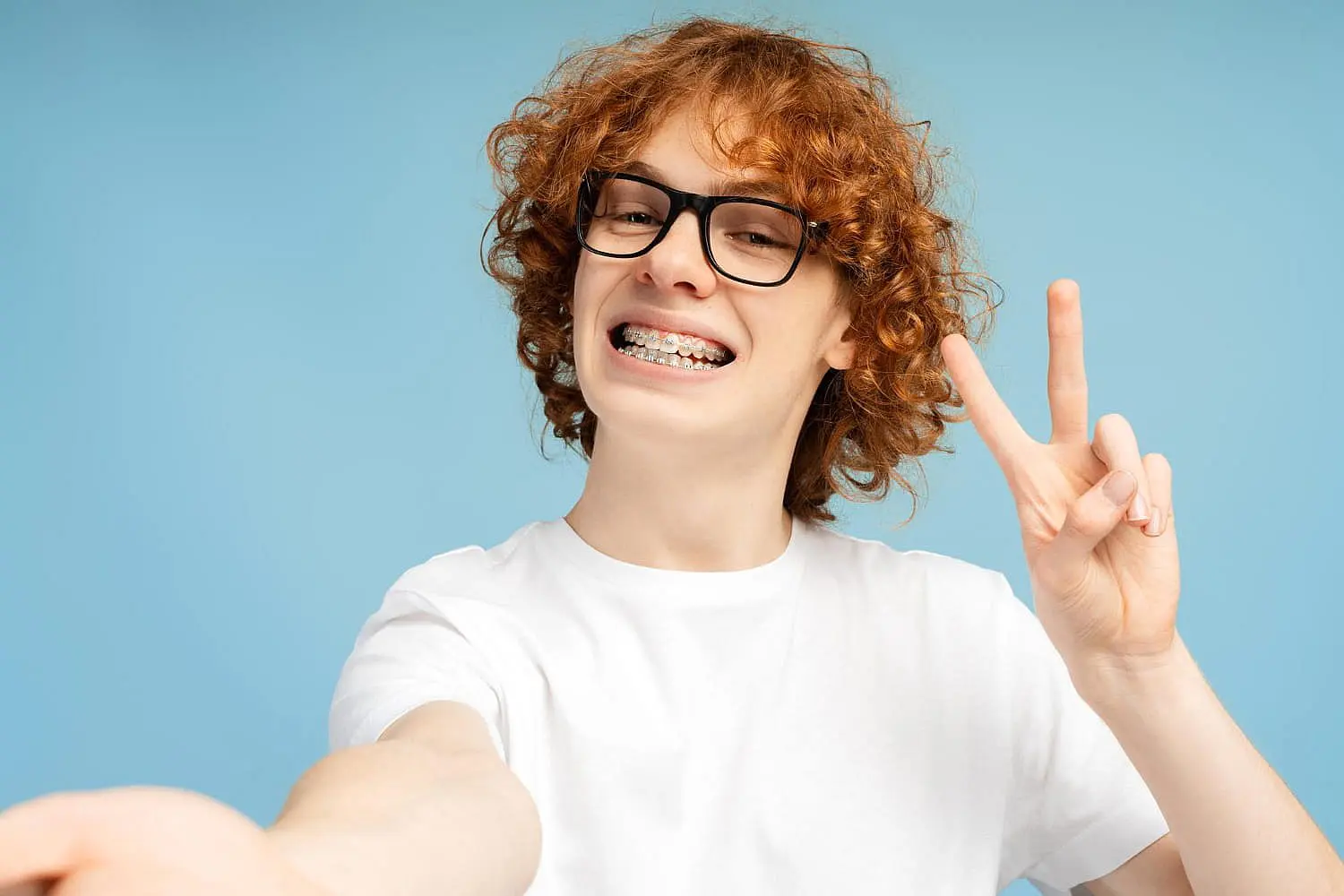 A cheerful teen with curly hair and glasses flashes a bright smile, proudly showing braces, as they form a peace sign against a calming blue background in Hamilton, OH