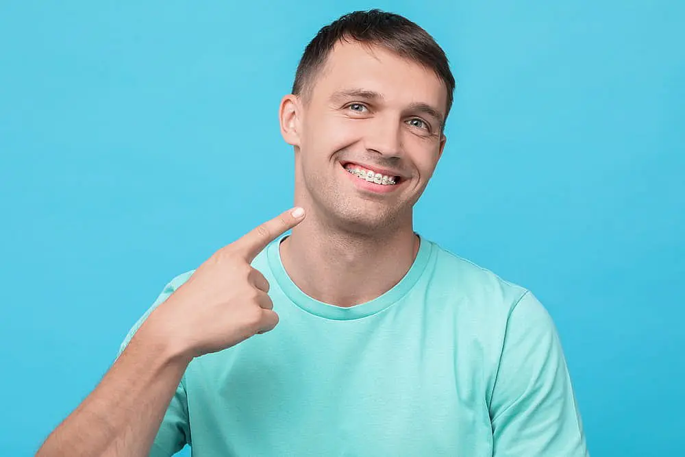 A man beams with confidence as he proudly points to his metal braces, dressed in a light blue shirt against a serene blue background in Liberty Township OH
