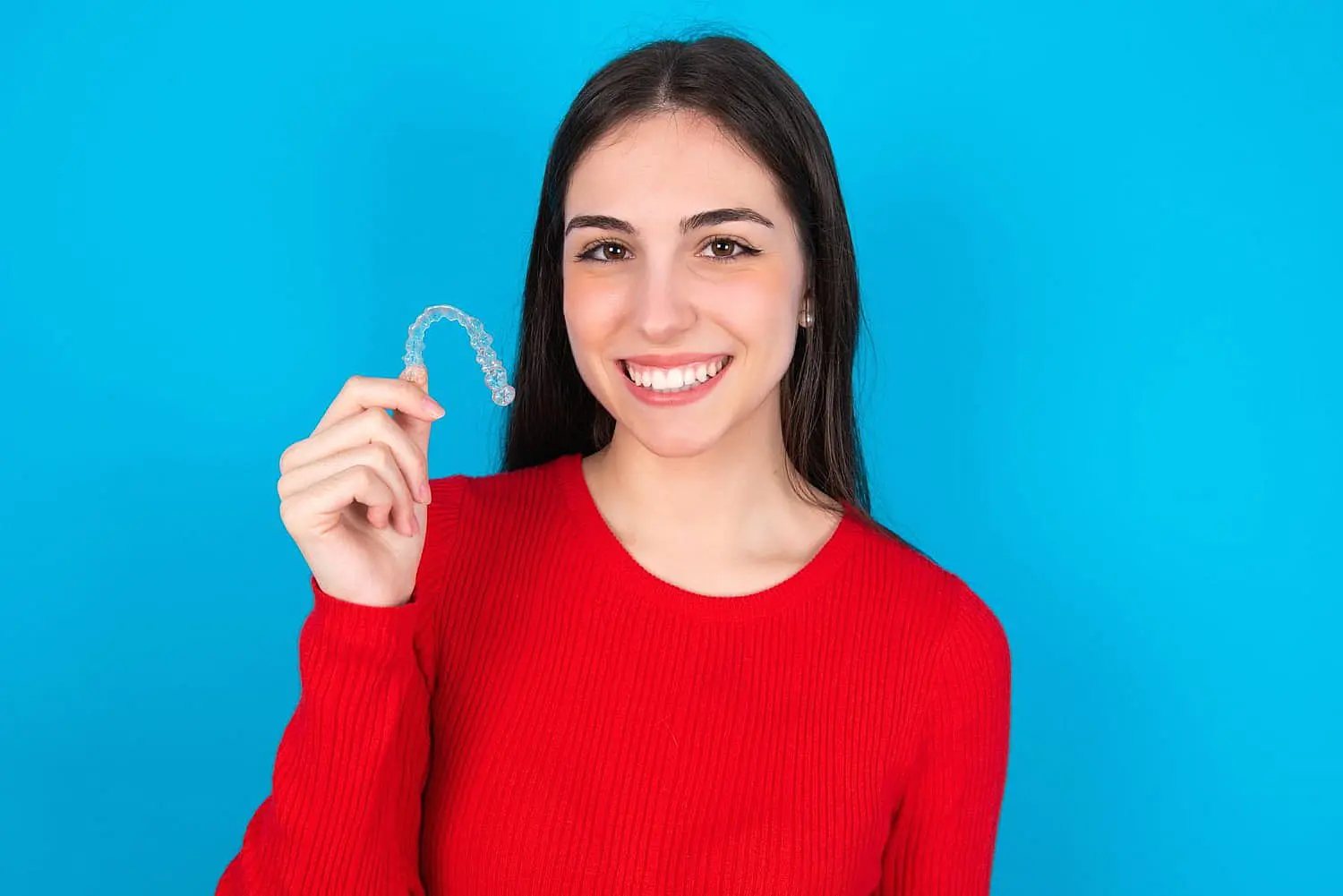 A woman in a red sweater smiles as she proudly displays her Invisalign clear dental aligner against a vibrant blue background in Liberty Township, OH