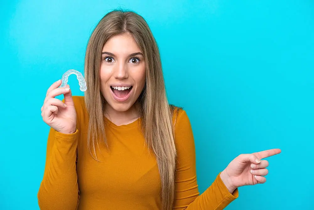 Woman in a mustard sweater holds an Invisalign dental retainer and points to the right against a blue background in Hamilton, OH