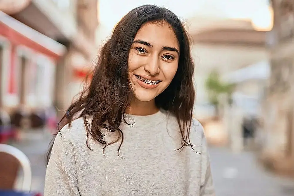 Young woman with long hair and metal braces smiling, wearing a gray sweater, standing outdoors with blurred buildings in the background in Liberty Township or Hamilton, OH