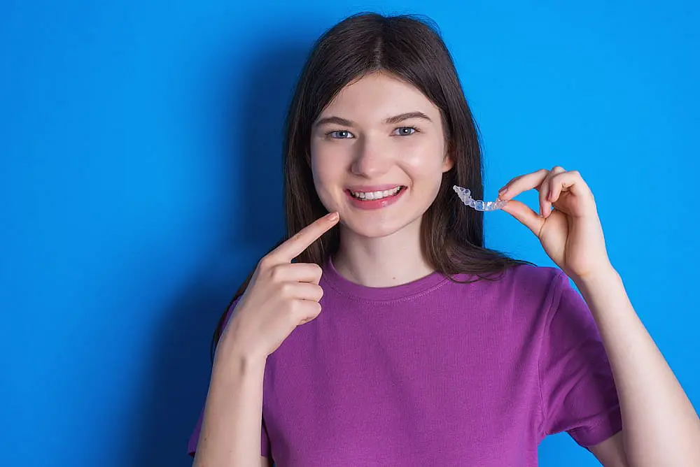 A person in a purple shirt smiles, showcasing their teeth while holding an Invisalign clear dental aligner against a blue background in Liberty Township, OH