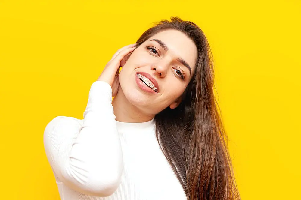 A person with long hair and clear braces, wearing a white top, touches their neck while smiling in front of a vibrant yellow background in Liberty Township OH