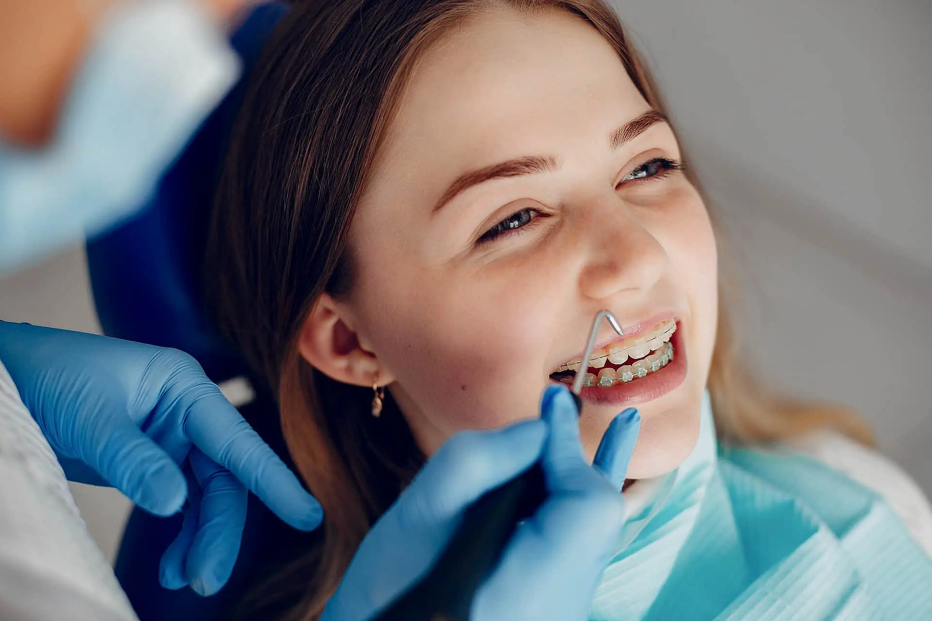 A dentist uses a tool to examine a patient with clear braces. The patient is smiling while seated in a dental chair, wearing a protective bib. The dentist's hands are gloved in Liberty Township, OH