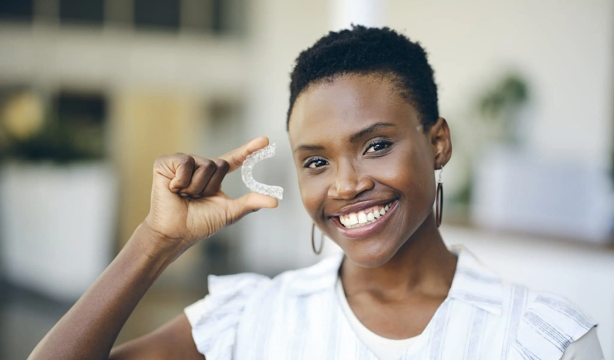 A person is smiling brightly, holding their Invisalign clear dental aligner while standing indoors in Liberty Township, OH