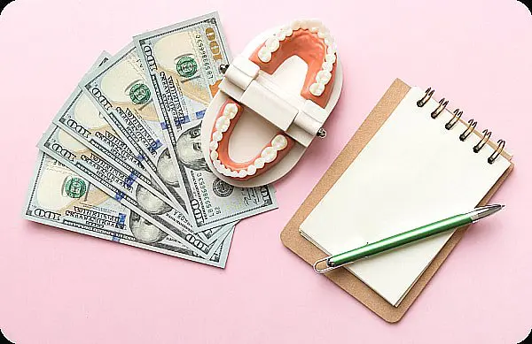 A set of artificial teeth with metal braces sits alongside dollar bills, a notepad, and a green pen on a vibrant pink background in Liberty Township or Hamilton, OH