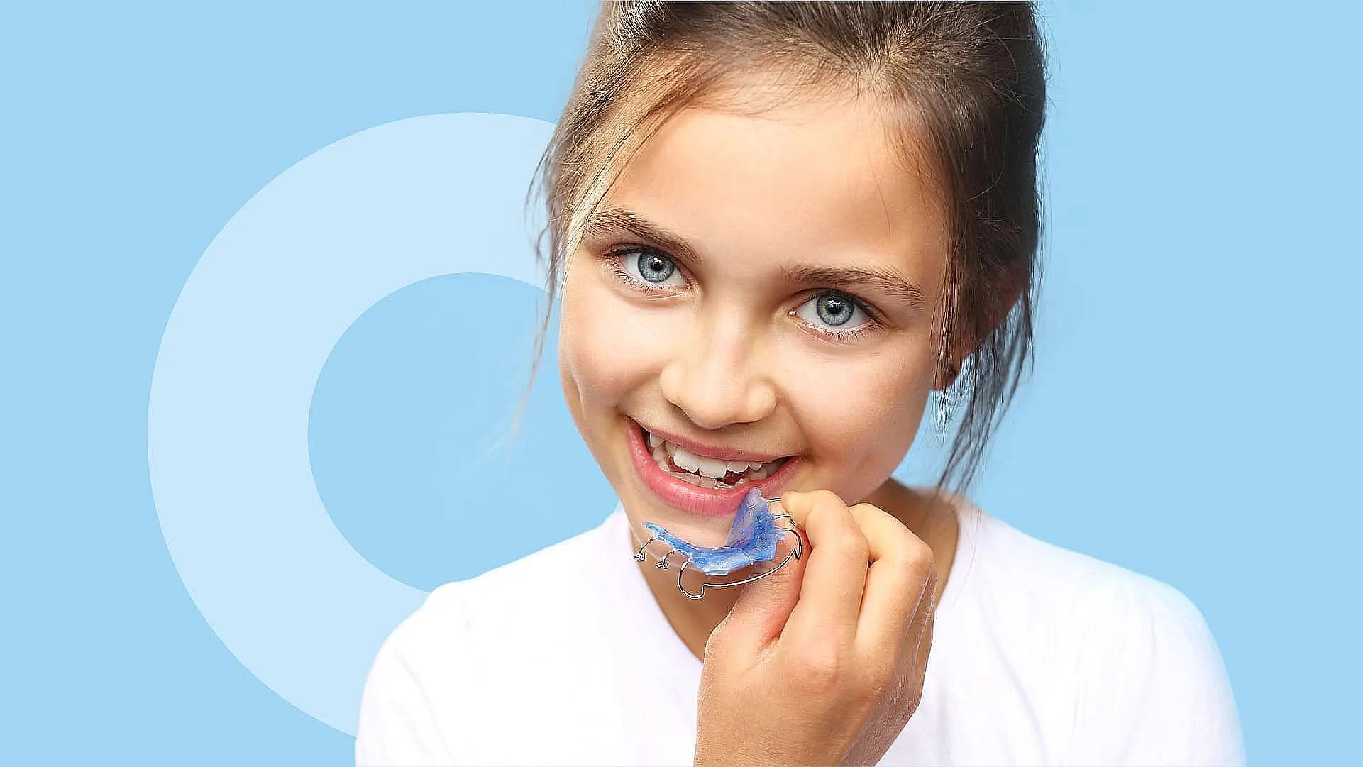A girl with light brown hair delicately holds an orthodontic appliance, her dental retainer, near her mouth against a soothing blue background in Liberty Township or Hamilton, OH