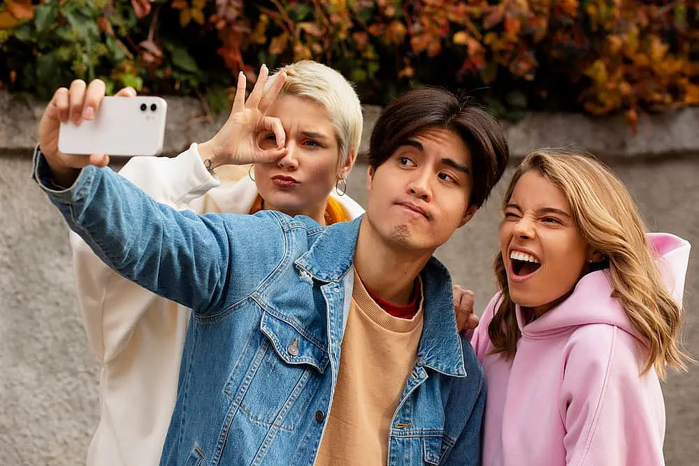 Three friends capture a selfie outdoors; one makes a hand gesture, another looks puzzled, and the third smiles widely. Amidst the autumn leaves, their casual attire hints at carefree days—like choosing between different types of braces for a unique fit in Liberty Township or Hamilton, OH