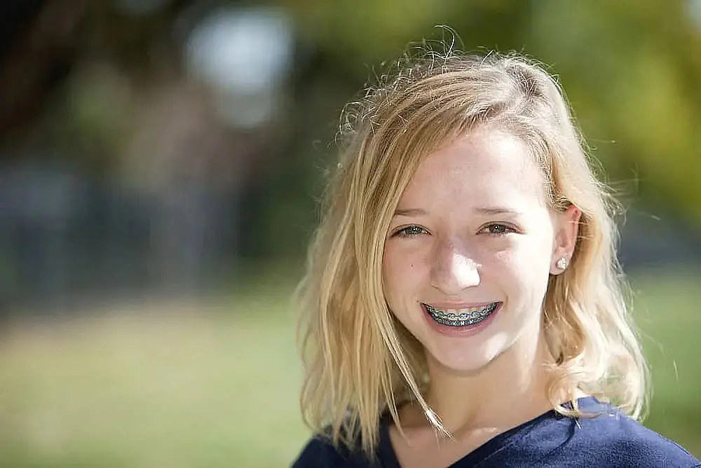 A young person with blonde hair beams outdoors, showcasing their braces and a navy top against a blurred natural background, confident in their smile thanks to an easy braces payment plan in Liberty Township or Hamilton, OH