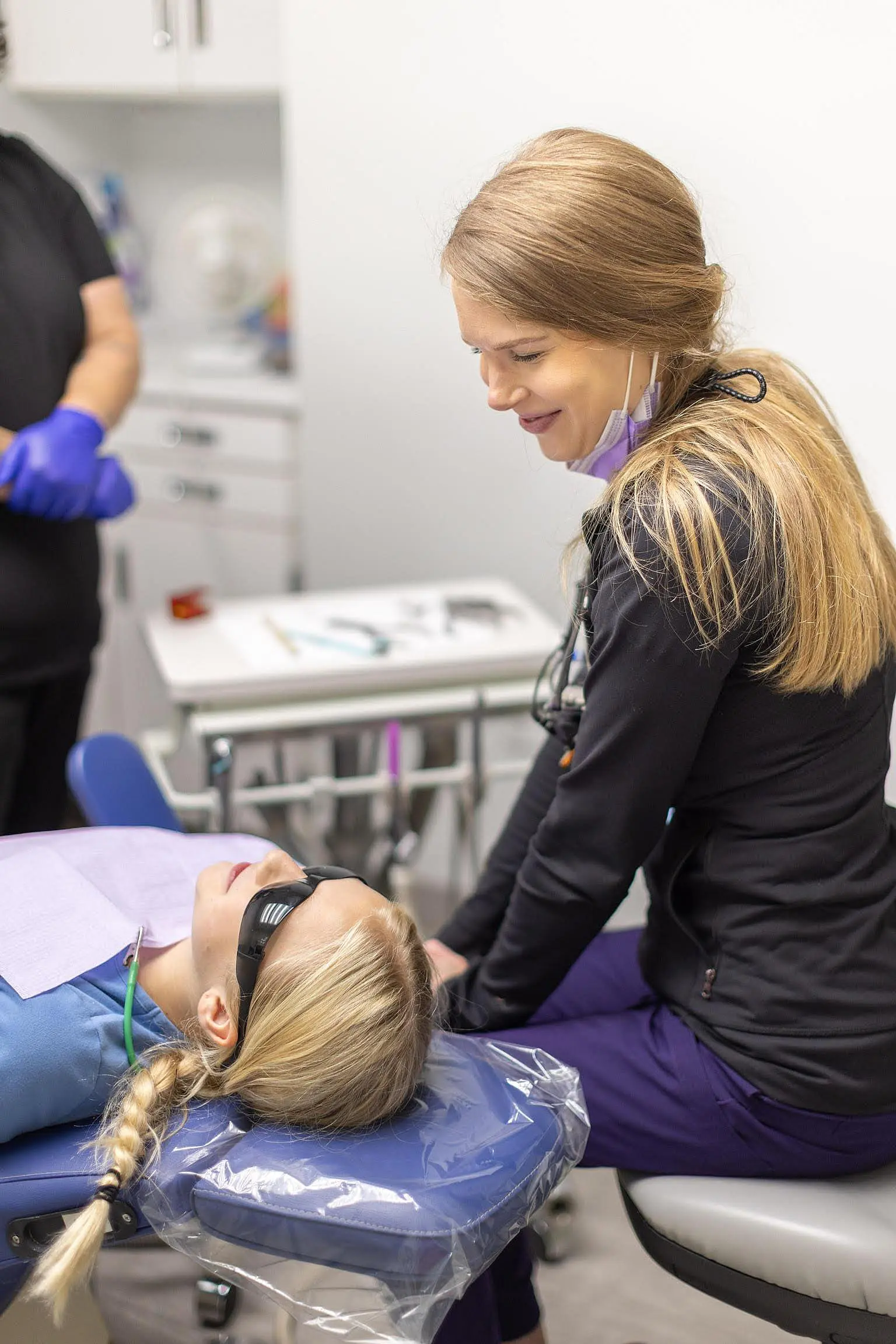 A dental professional speaks with a patient lying on a dental chair, both with long blond hair, in a bright clinic room. Another person stands in the background at Queen City Orthodontics in Hamilton, OH.